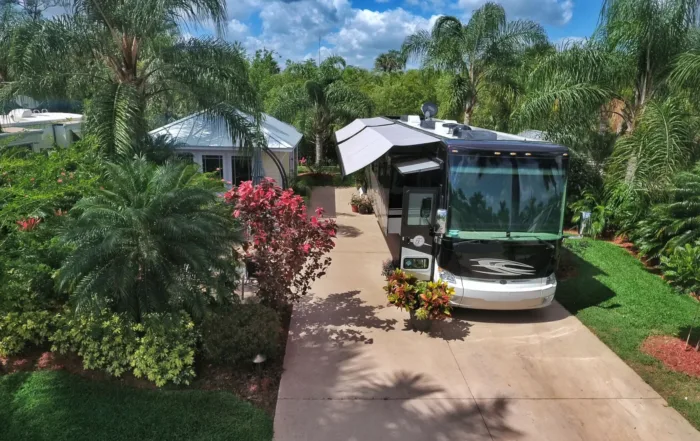 Luxury motorhome parked on a landscaped RV lot with palm trees, patio, and a small pavilion under a blue sky.