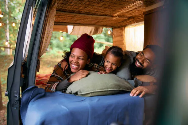 Smiling family relaxing on a bed inside a camper van at a wooded campsite with string lights.